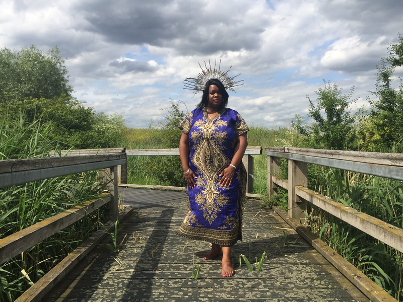 A black woman standing majestically on a wooden bridge among reeds and marshland. She is wearing a headdress and a decorative purple and gold dress.