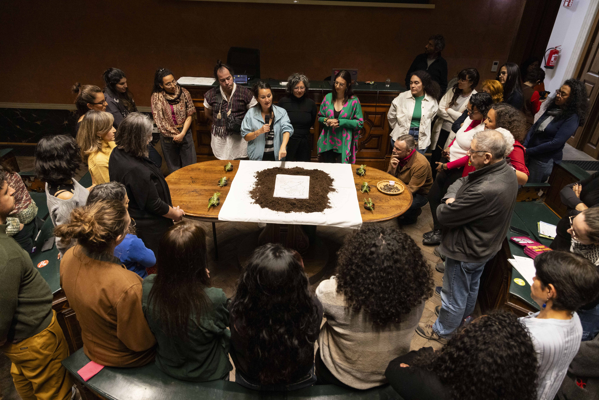 A group of approximately 30 people is standing around a table in the Anatomy Hall of the Academy of Fine Arts Vienna. One person is speaking into a microphone and pointing to soil that is placed on the table.
