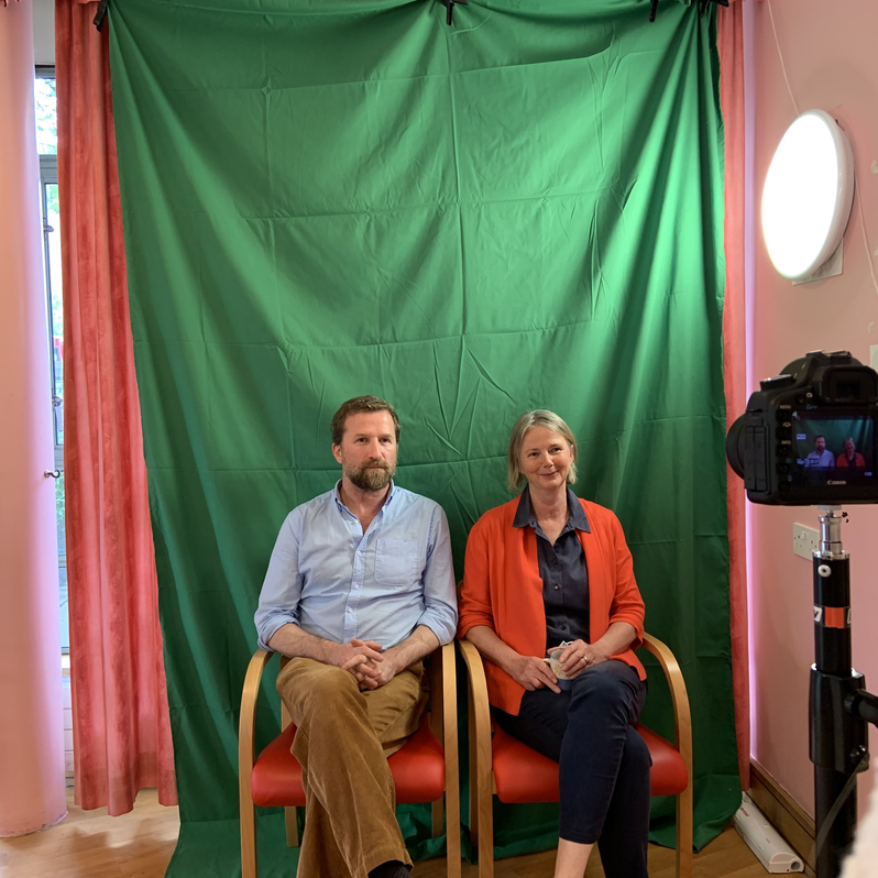 A white man and woman sit in front of a green screen with a DLSR camera filming them