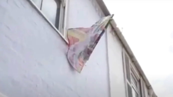 A video still showing a flag attached to the outside of a house. The flag is multicoloured and collaged.