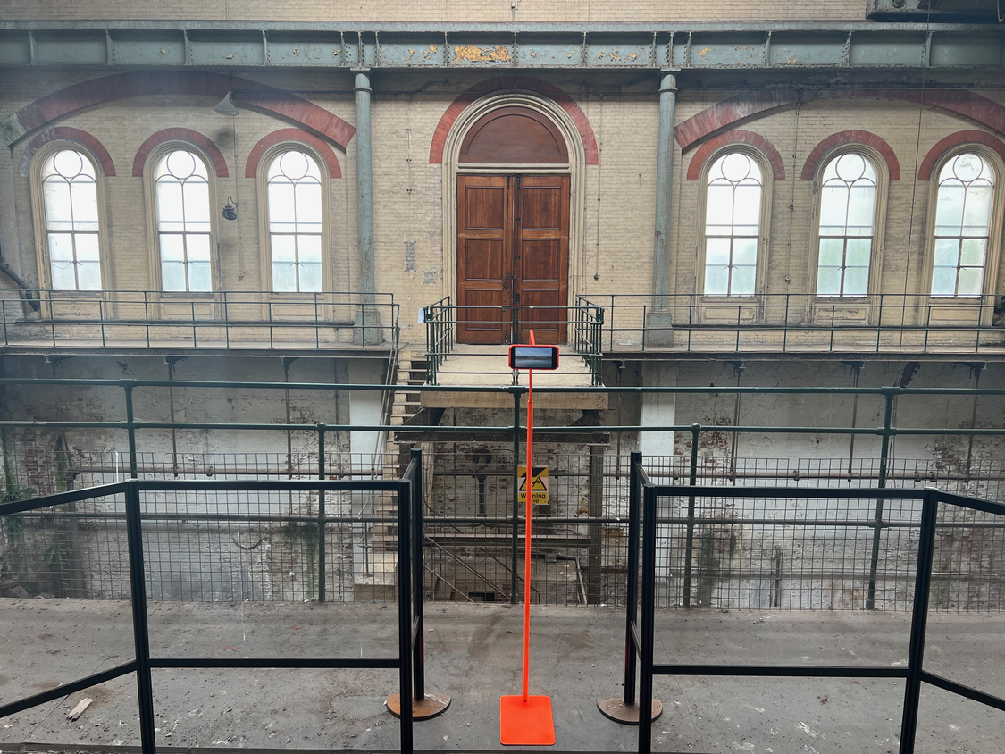 a florrstanding mobile phone stand in hi-vis orange, with orange cables trailing off, a mobile phone with a video still image on it with a background behind of the triple expansion house at Crossness