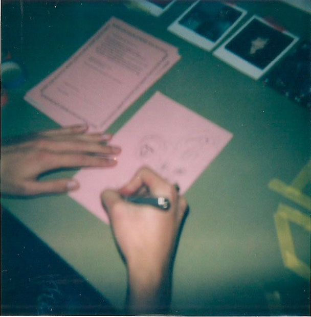 polaroid photo of someones hands writing on a pink piece of paper.
