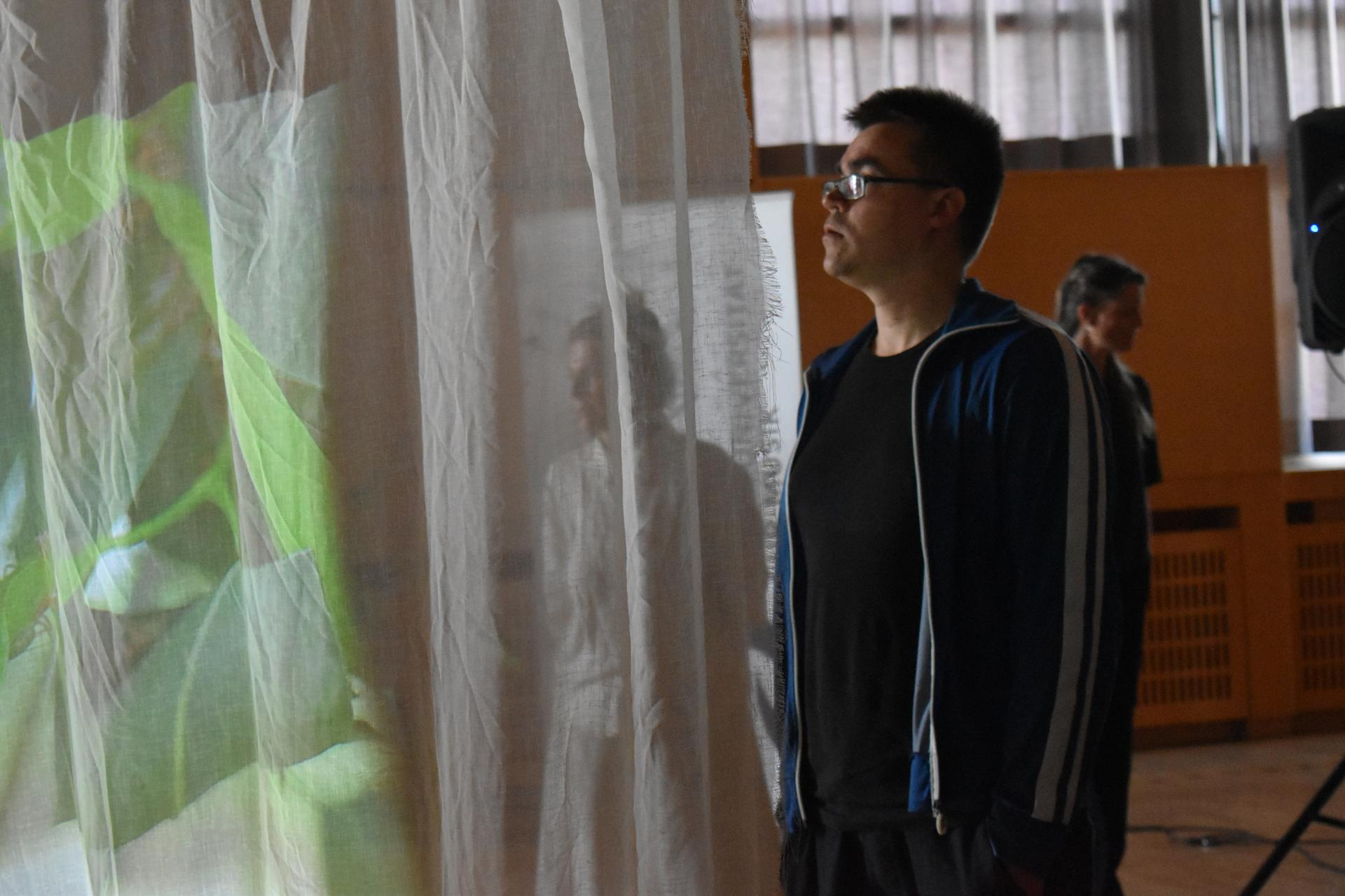 On the right, a person pauses to listen to the quadrophonic sound installation during the lecture-performance. On the left we see a detail of leaves projected on a semi-opaque curtain.