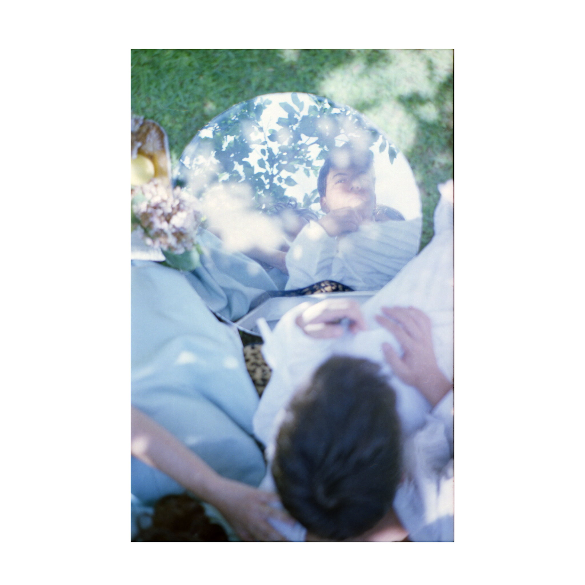 The photograph shows a circular mirror placed outdoors on grass, reflecting the face of a young person in white clothing, along with branches and leaves above them. Around the mirror, parts of other figures are visible, including an arm, a hand, and folds of light blue and white fabric. On the left edge of the frame, a tray with lemons and flowers can be seen. The overall lighting is bright, with soft dappled natural daylight illuminating the scene.