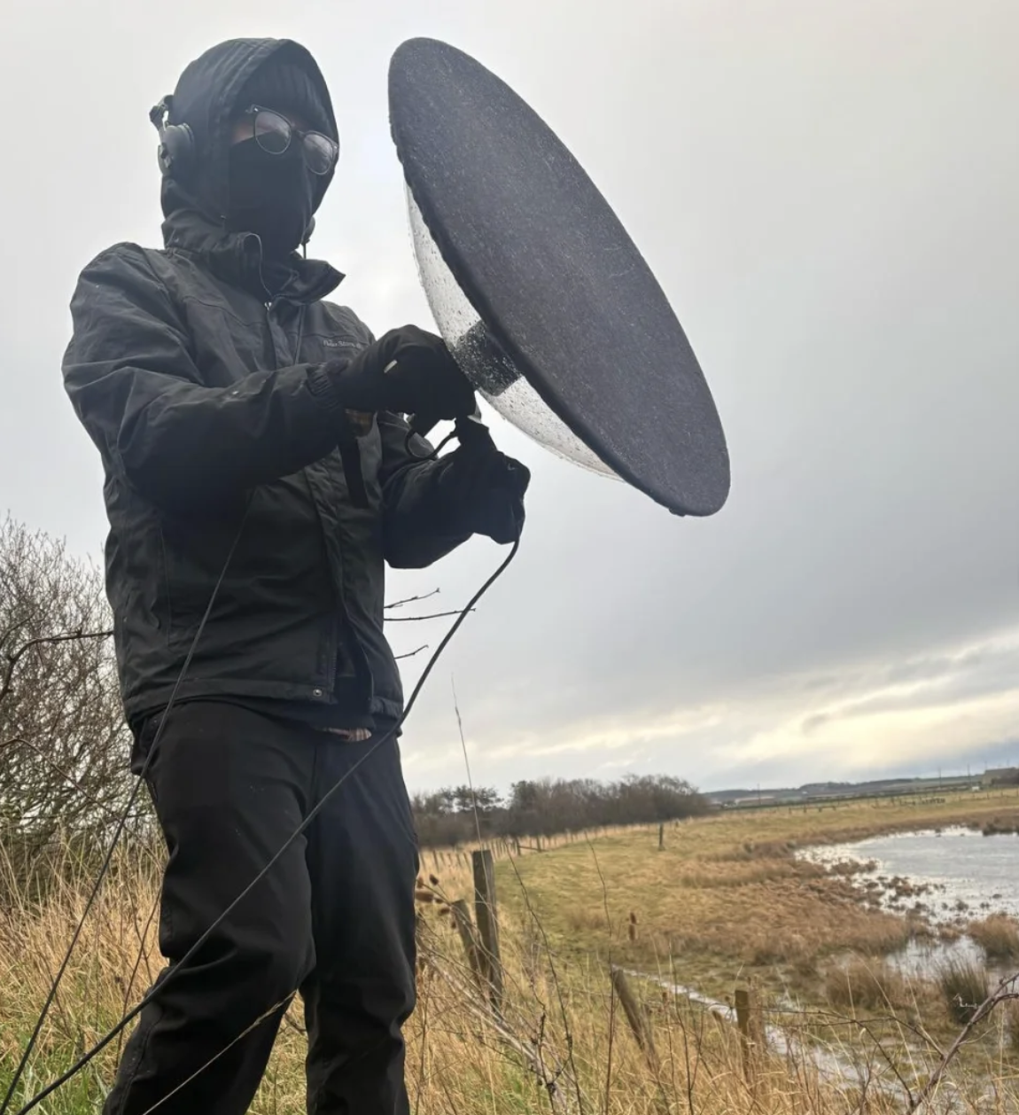 A outdoor photo of Jorge Boehringer holding a parabolic reflector with a microphone inside. Boehringer is wearing all black and wrapped up against the cold.