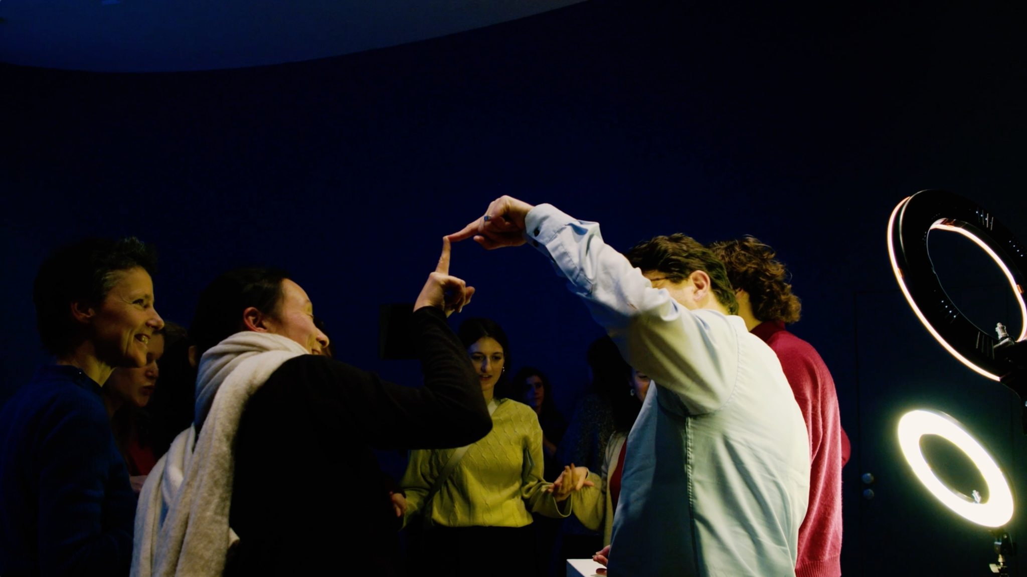 A group of people stand together in a dimly lit room with a blue background. Two people in the foreground are touching their index fingers to form an arch, smiling and interacting playfully. Others watch and smile nearby, with circular studio lights illuminating the scene.