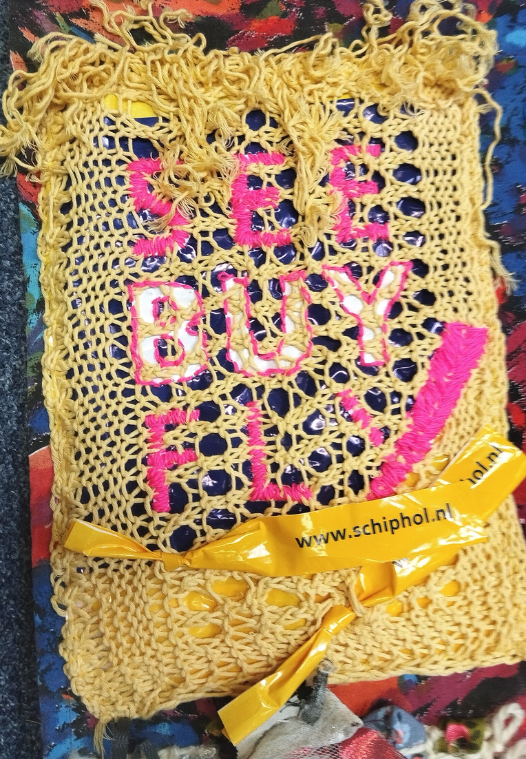 "See, Buy, Fly. The Web of Life is Torn by Consumerism."  Materials: netting knitted with essentials Organic Cotton by Rico, mustard colorway, color 003; plastic bag from Schiphol Airport Duty Free Shop, and 100% acrylic yarn from ON line Linie 50, Prima, color 0033 used for embroidery.