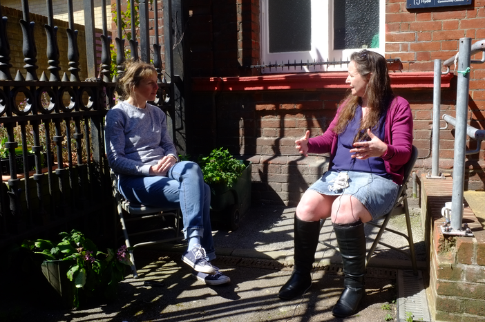 A photo of two women sat outside on chairs outside a red brick building and some black railings.