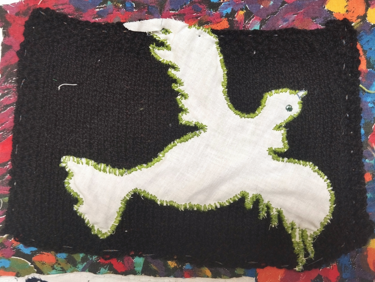 Knitted background made of black wool from maker Space stash in stockinette stitch with moss stitch border, application made from gifted cotton fabric, dove cut out and stitched on with embroidery floss in green.